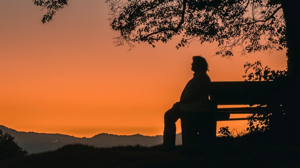 an aged lady waiting on bench in front of a beautiful sunset